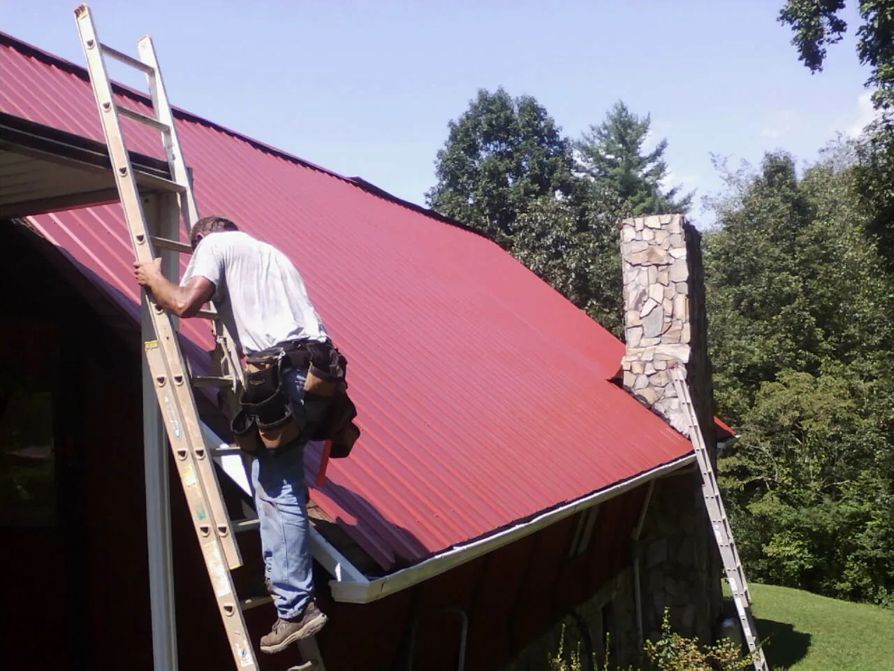 Roofer on a ladder beside a red metal roof and stone chimney — Soffit & Fascia Repair by Roofing Brothers in Culloden